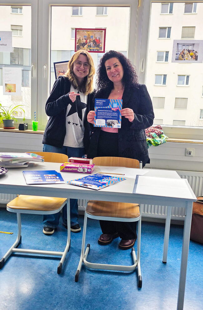 Zwei Frauen stehen lächelnd an einem Tisch im Sprachinstitut Böblingen und halten ein Informationsheft in der Hand.