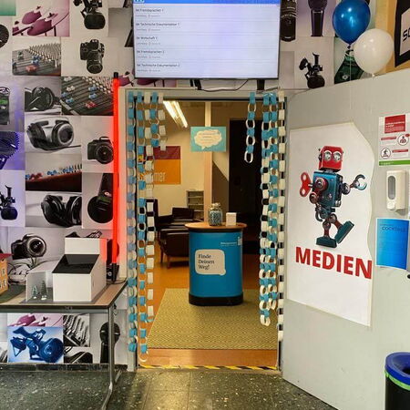 Ein bunt dekorierter Eingang mit Kameraplakaten und einem Schild mit der Aufschrift "Medien" führt zu einem Empfangsbereich.