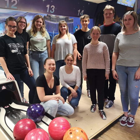 Eine Gruppe junger Menschen posiert lächelnd in einer Bowlinghalle vor den Bahnen mit bunten Bowlingkugeln.