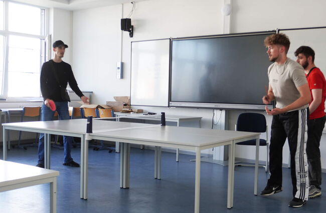 Drei Schüler spielen Tischtennis in einem Klassenraum, während ein Monitor im Hintergrund sichtbar ist.