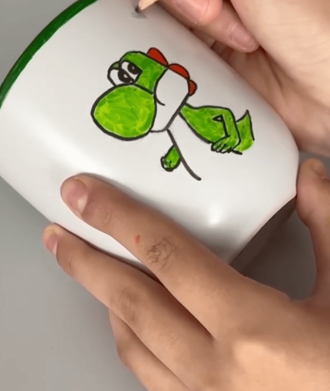 Ein Kind malt mit einem Bleistift eine grüne Figur auf eine weiße Tasse während des Projekttags.