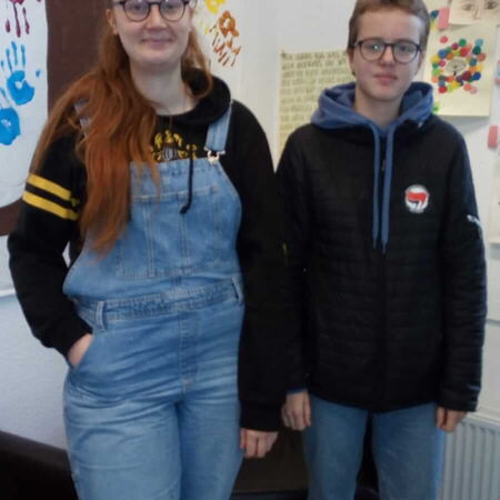 Zwei Personen stehen lächelnd vor einer Wand mit bunten Handabdrücken und Zeichnungen in einem kreativen Raum.