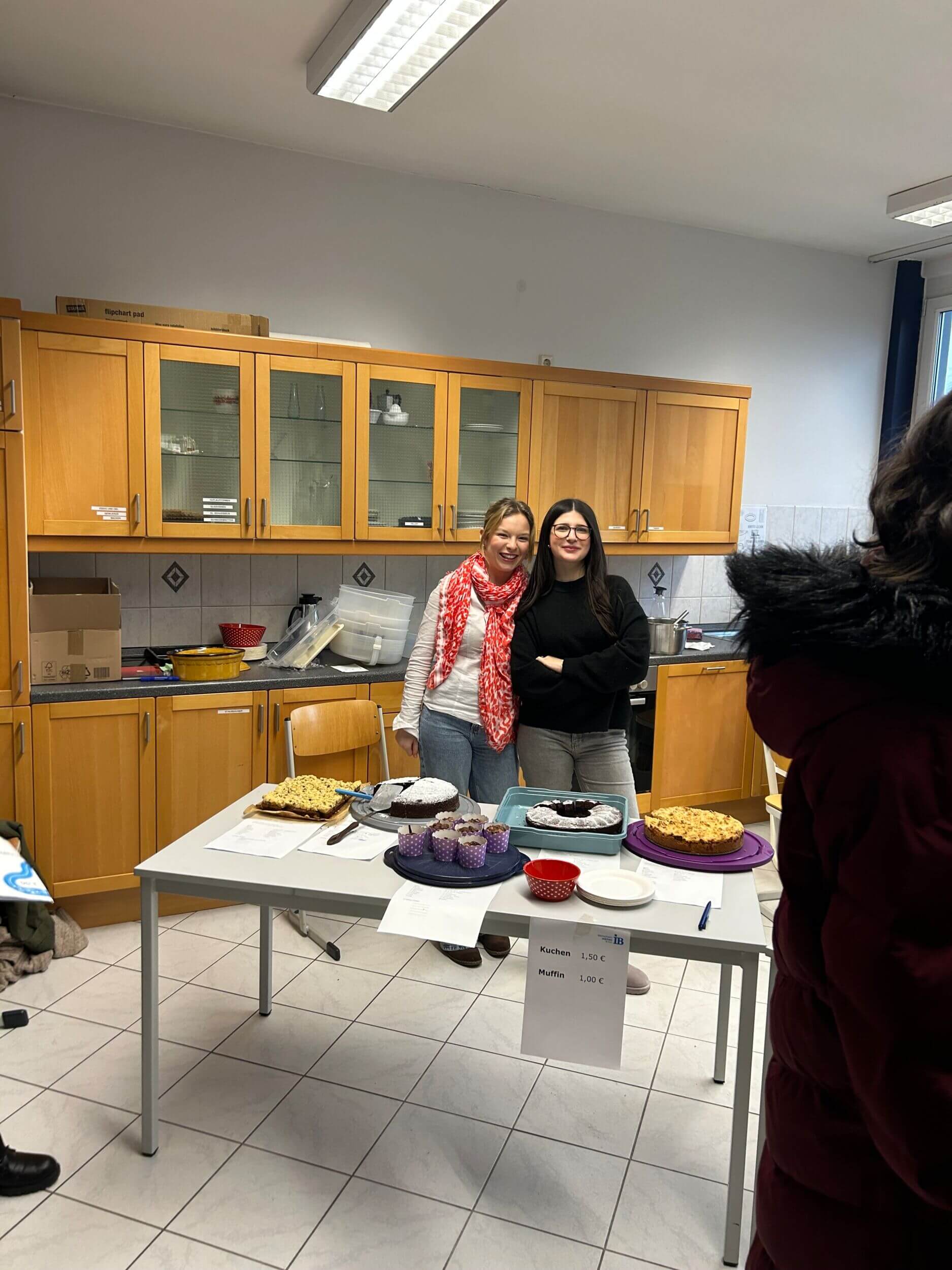Zwei Frauen stehen lächelnd hinter einem Tisch mit Kuchen und Muffins in einer Küche, bereit für einen Kuchenverkauf.